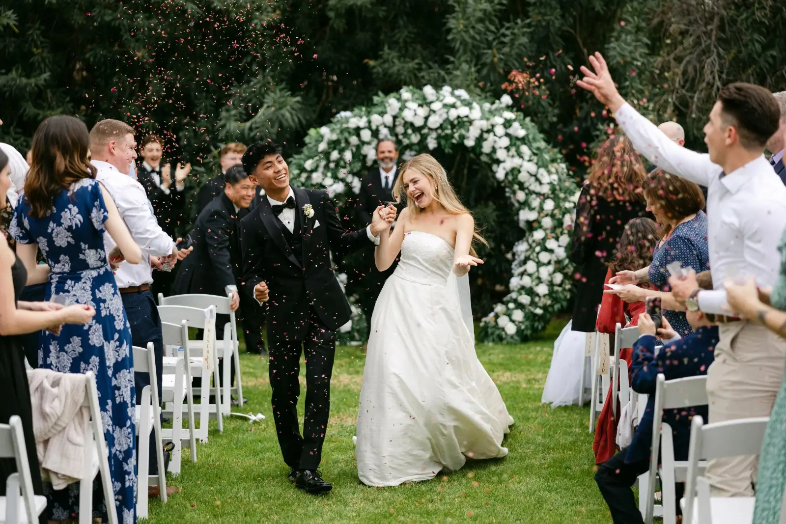 Couple walking back up the aisle in front of a white rose arch as guests cheer